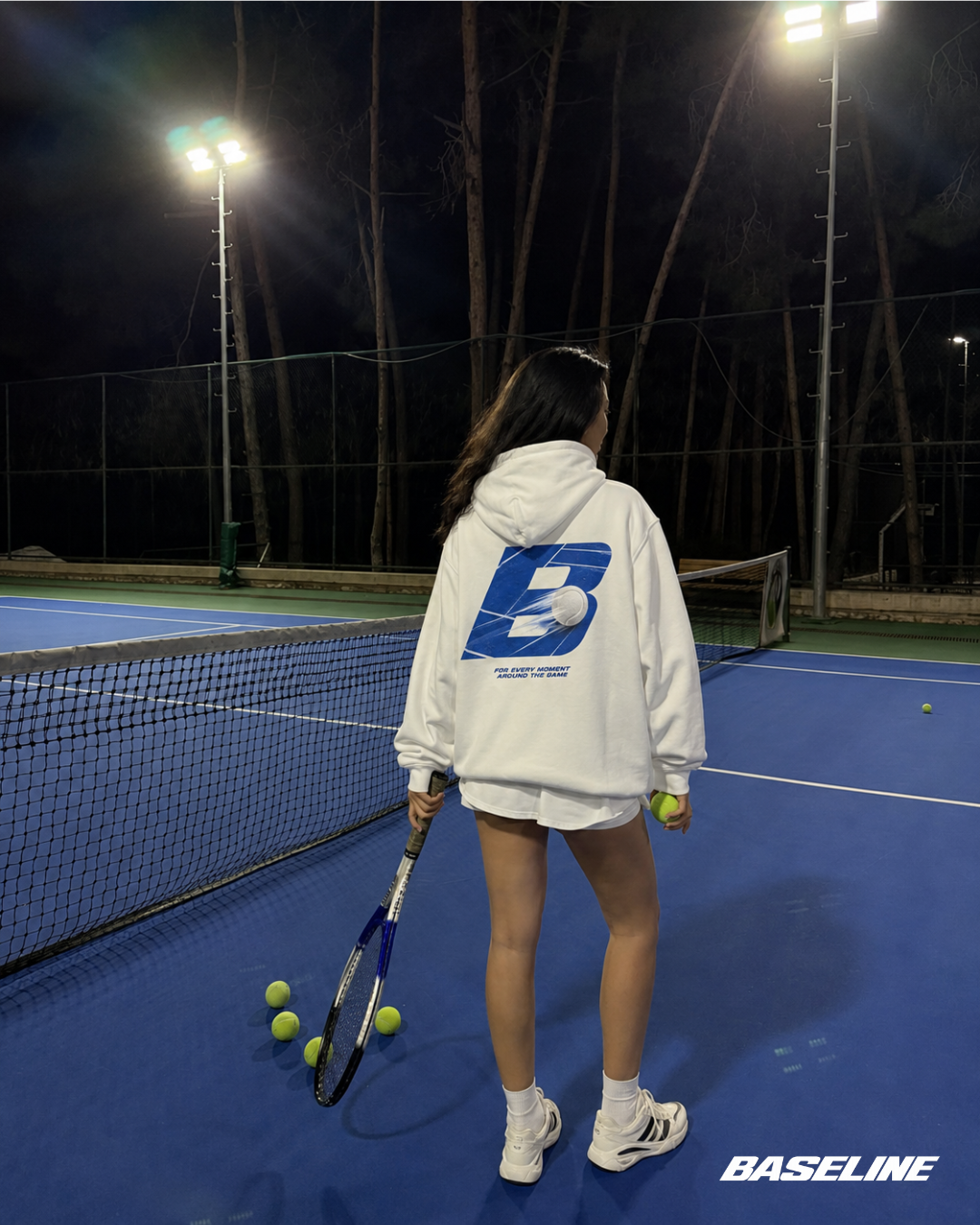 Baseline Court Blue hoodie worn beside a tennis court